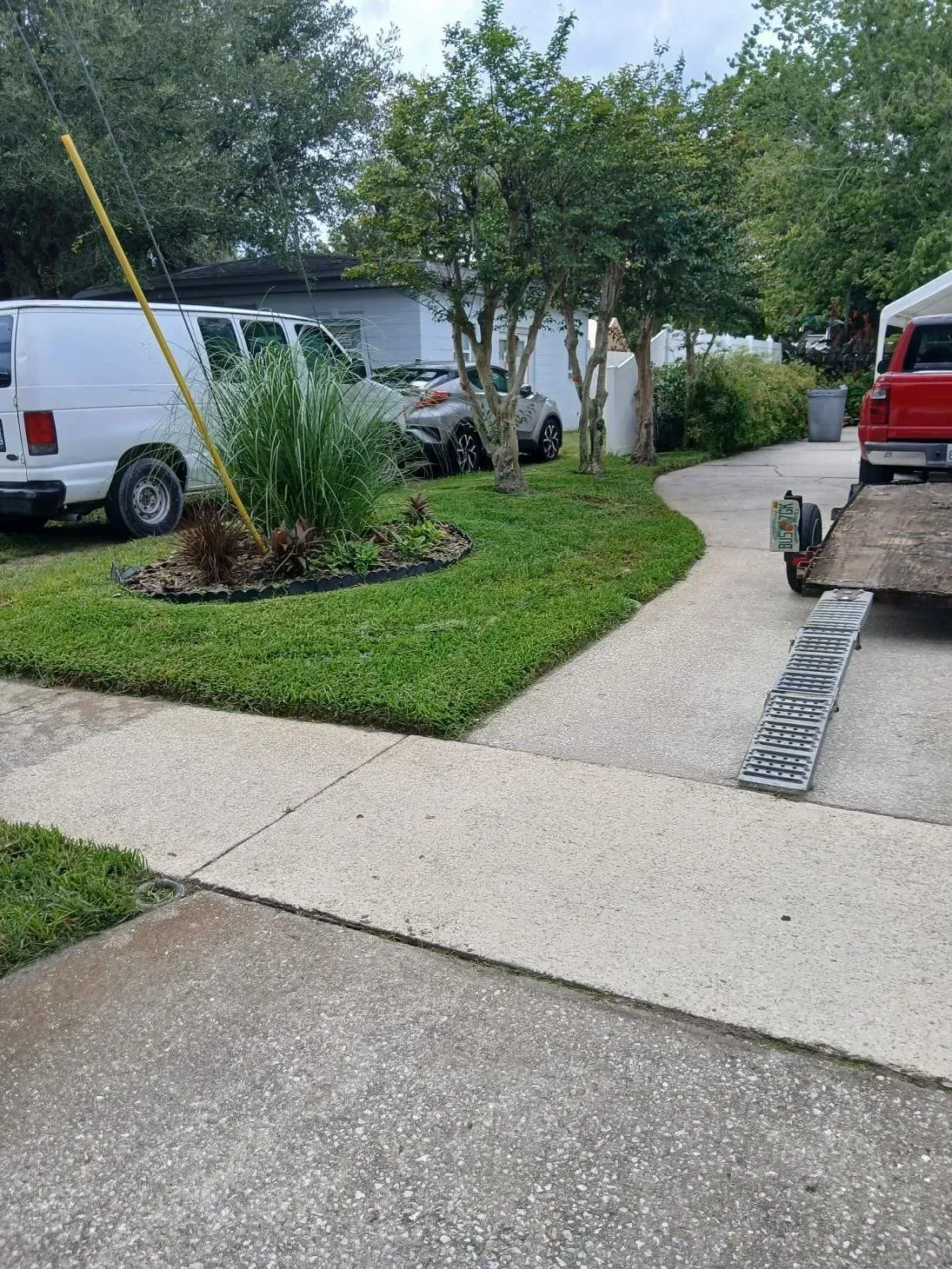 Residential driveway and walkway beside a lawn and landscaped bed, with parked white van and red truck.