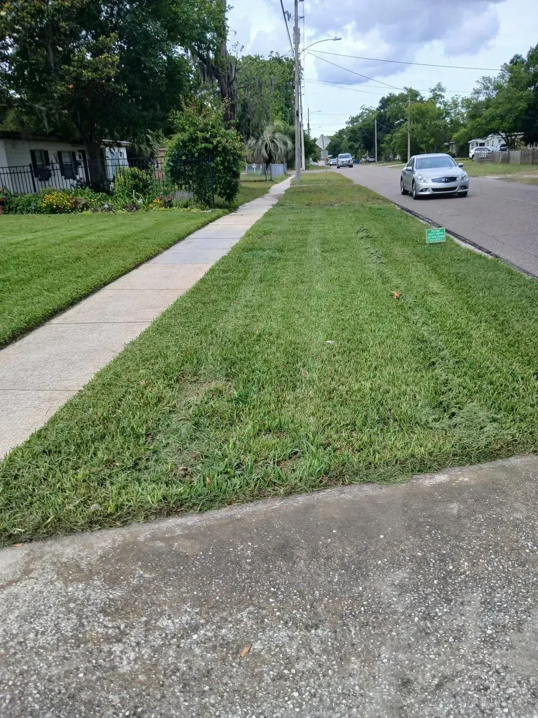 Residential street with grassy verge, sidewalk, parked silver car, and cloudy sky.