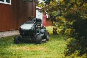 Black riding lawn mower on grass beside a red building, partly framed by bushes
