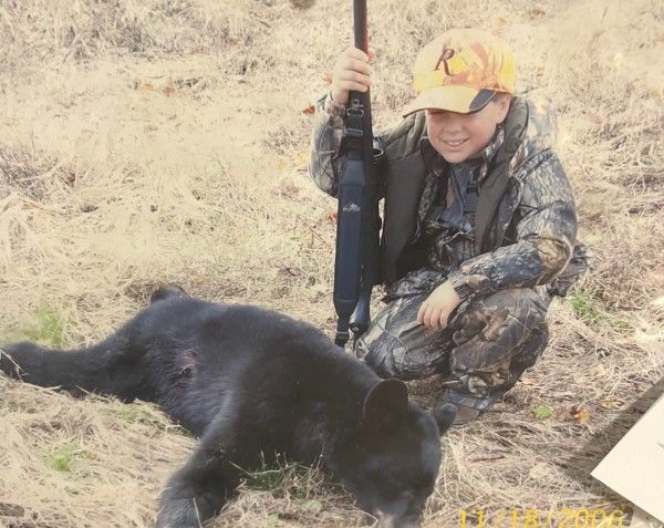 Hunter kneels beside a black bear in dry grass, holding a rifle upright and smiling.