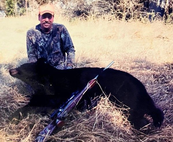 Hunter kneeling beside a black bear in dry grass, with a rifle in the foreground.