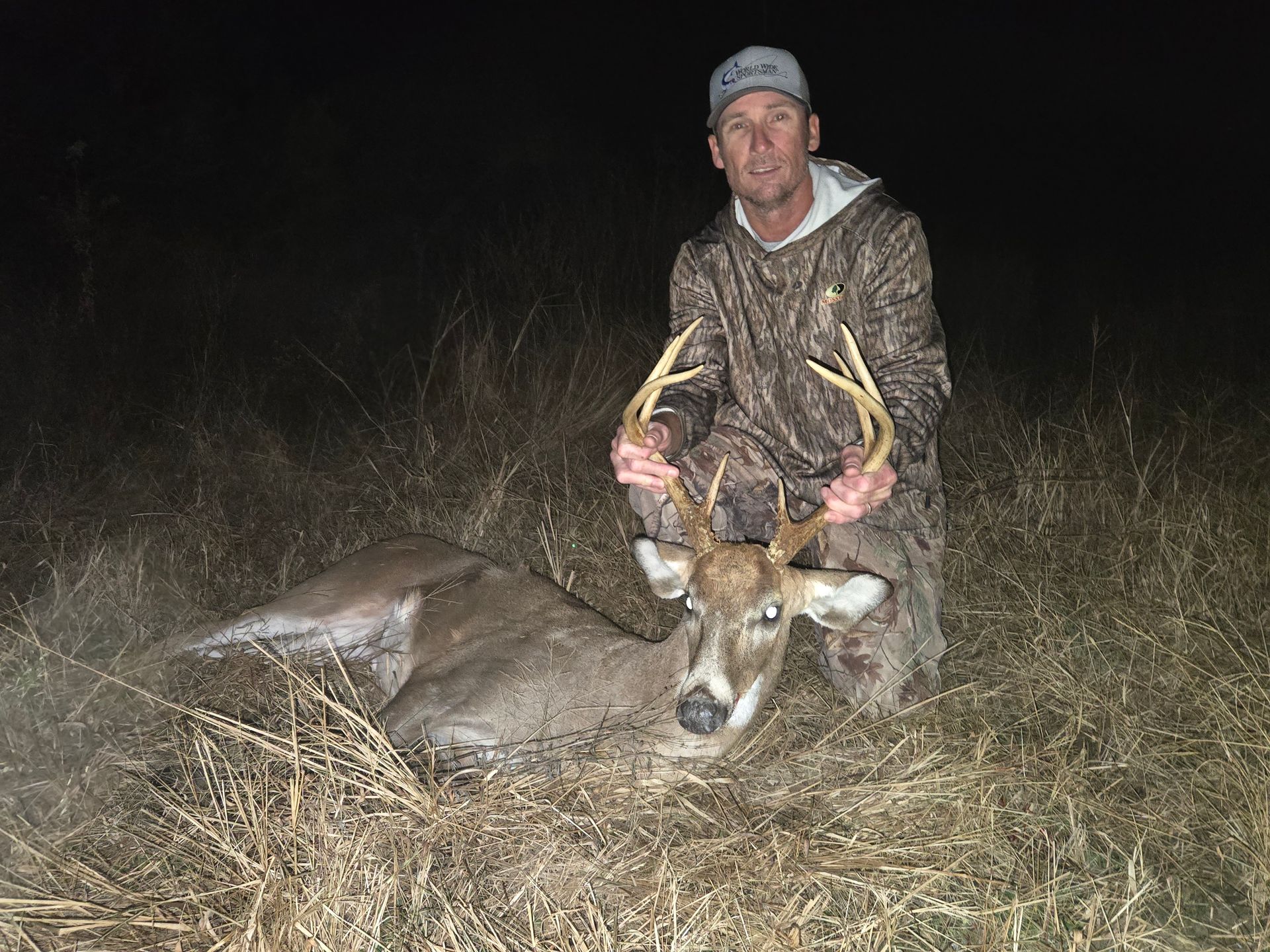 Man kneels beside a large deer with antlers in tall grass at night, holding the antlers.