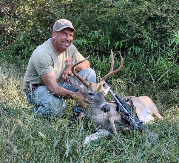 Man kneeling in grass beside a hunted deer with antlers, holding it in a wooded field.