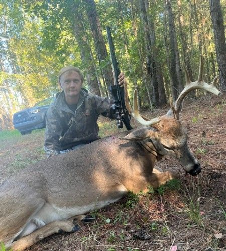 Hunter kneels beside a large deer in a wooded area, holding a bow.