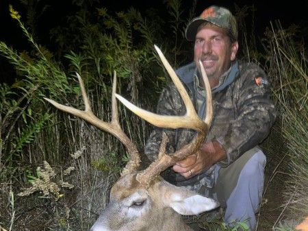 Hunter kneels beside a large deer at night in brush, holding its antlers and smiling.