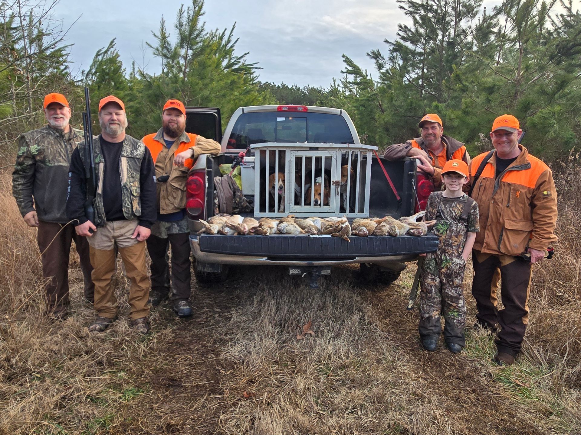 Hunters posing with harvested ducks beside a pickup truck in a field, wearing orange hats and camo gear