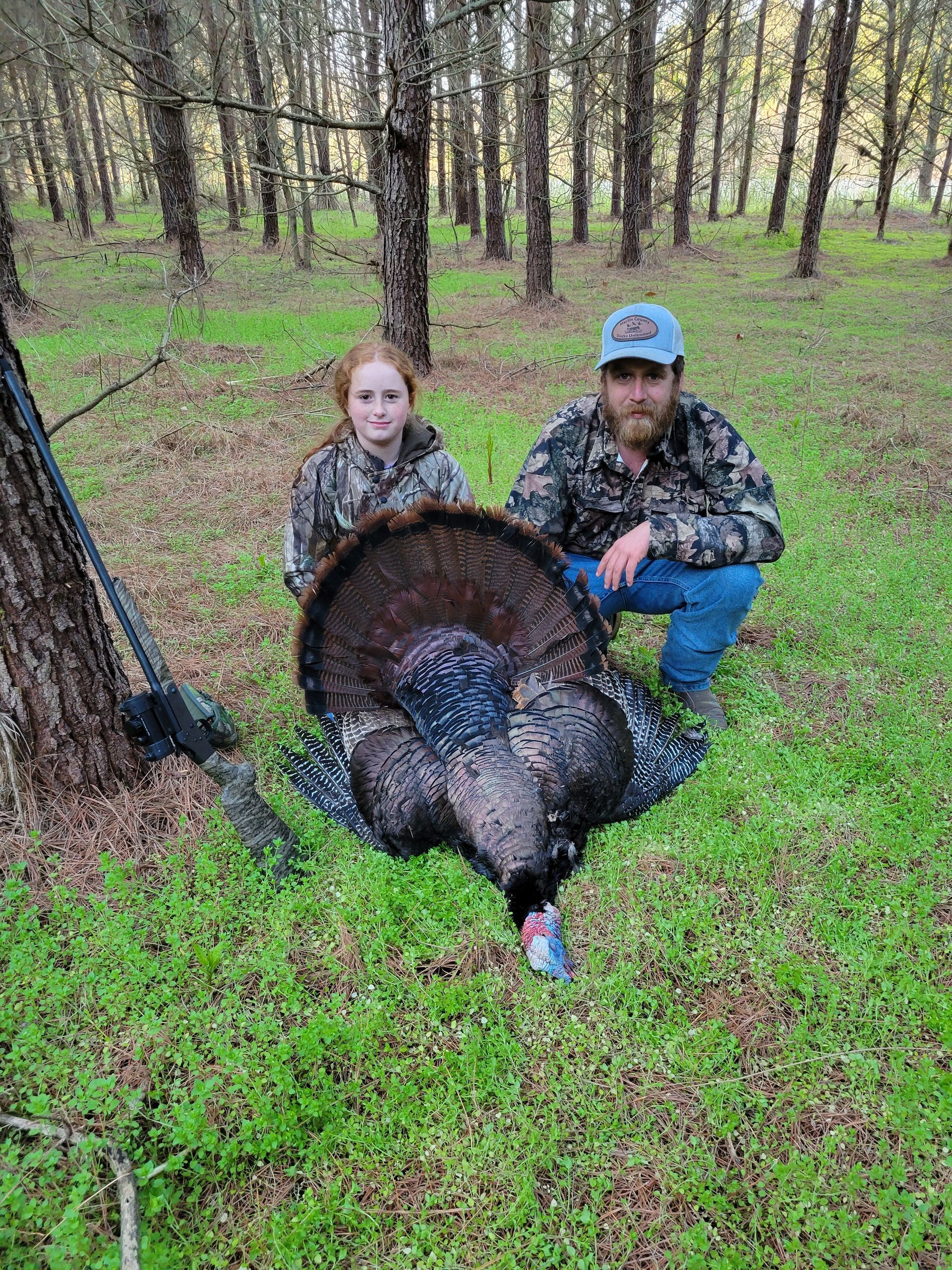 Camouflaged hunter kneels beside a child and a wild turkey in a forest clearing.