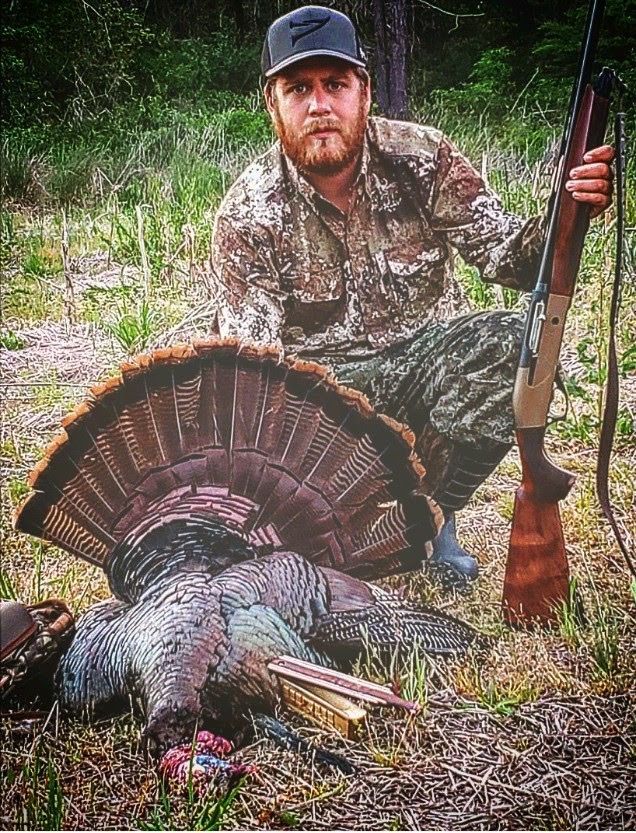 Hunter kneels beside a large wild turkey, holding a shotgun in a grassy wooded area.