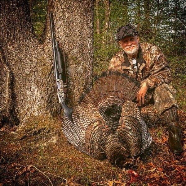Hunter kneels beside a large wild turkey in a wooded area, with a shotgun propped against a tree.