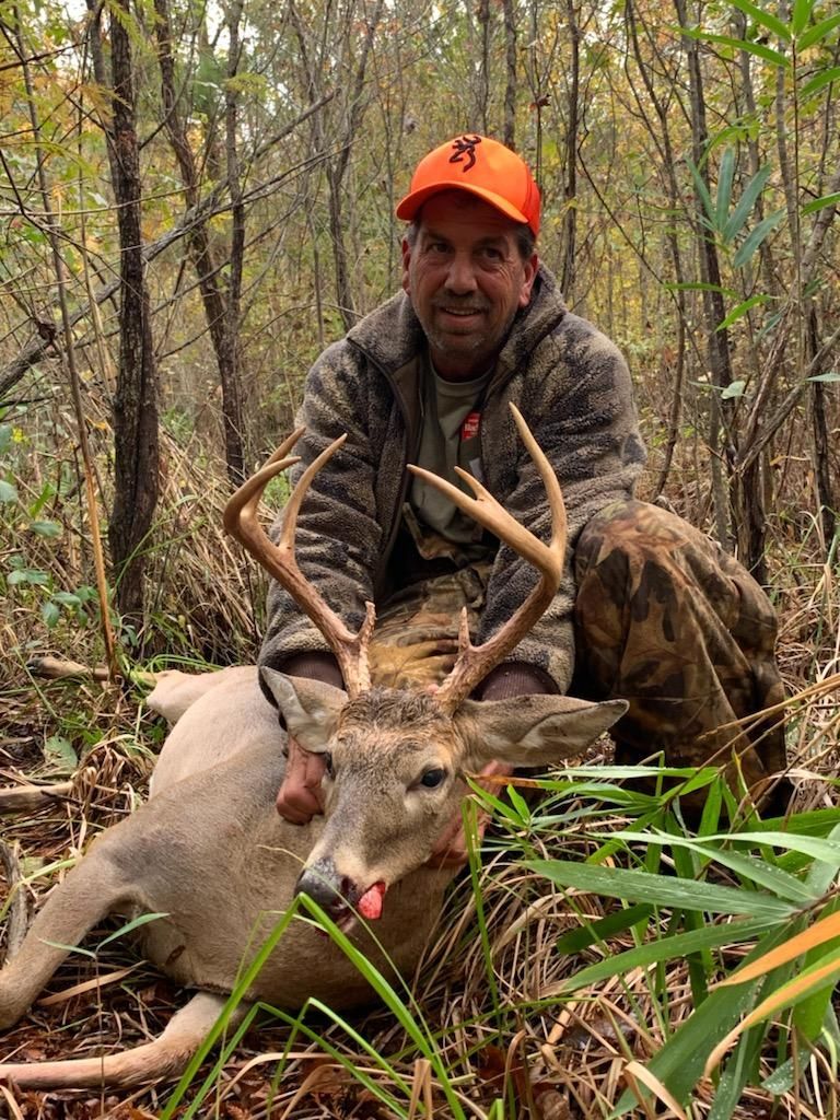 Hunter crouching behind a large buck in a wooded area, wearing camo and an orange cap.