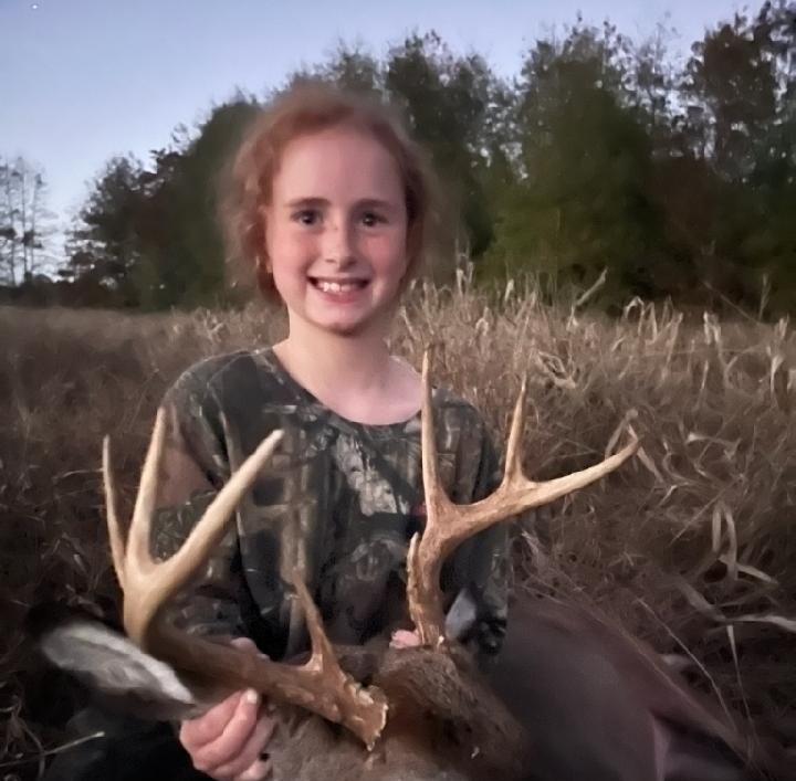 Smiling person in camouflage holding a deer antler in a grassy field at dusk