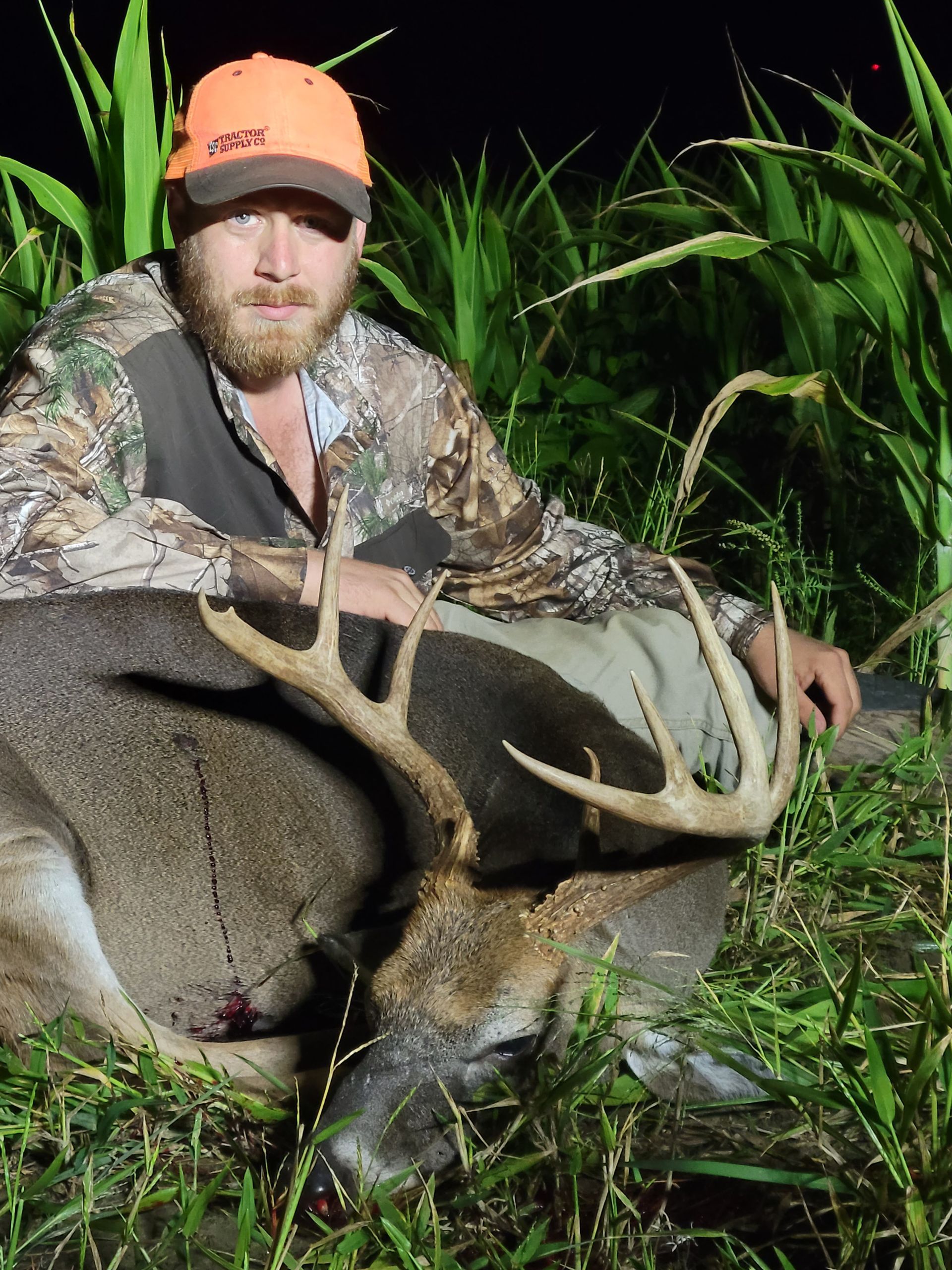 Hunter kneels beside a large deer in tall grass at night, wearing a camouflage jacket and orange cap.