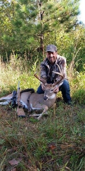 Hunter kneels beside a dead deer in tall grass, holding its antlers in a wooded area.