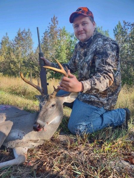 Man kneels beside a deer he has harvested in a grassy field, holding its antlers under a blue sky.