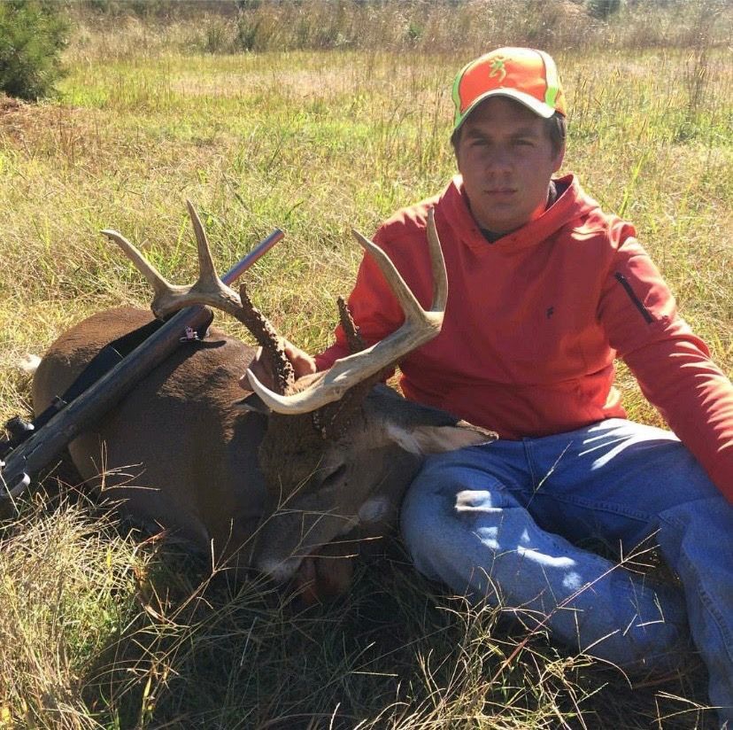 Hunter kneels beside a large deer in a grassy field, wearing a red jacket and orange cap.