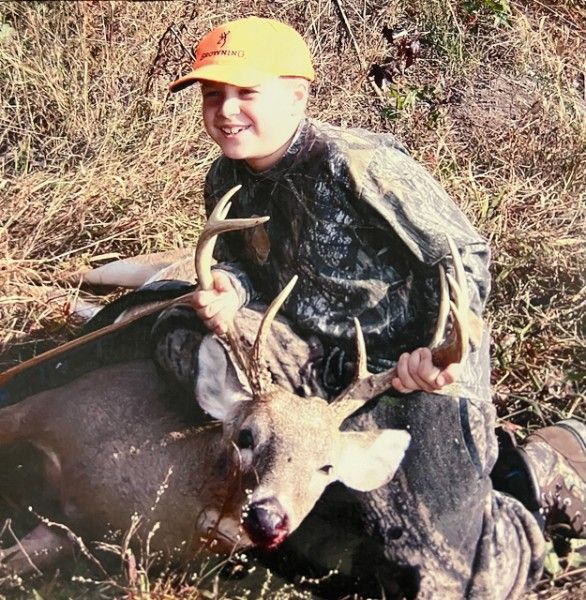 Hunter in orange cap kneels beside a deer with antlers in tall brush.