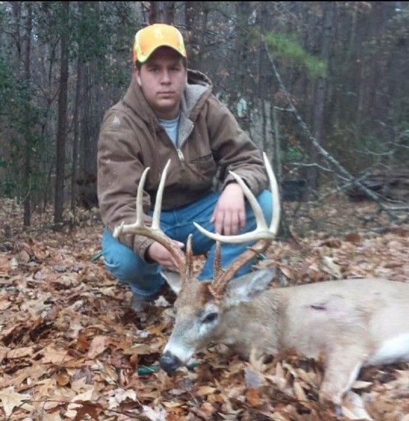 Hunter kneels beside a deer in the woods, wearing a yellow cap and brown jacket.