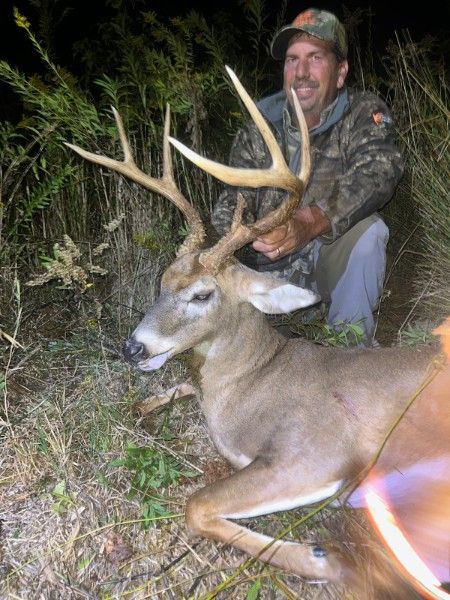 Hunter kneels beside a large buck with antlers in tall grass at night.