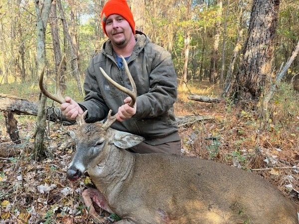 Hunter kneels beside a deer in a wooded area, holding the antlers and smiling, wearing an orange beanie.