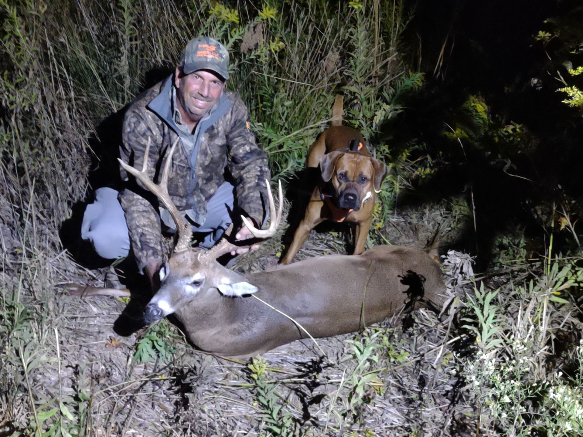 Hunter kneels beside a deer at night while a dog stands behind in brush.