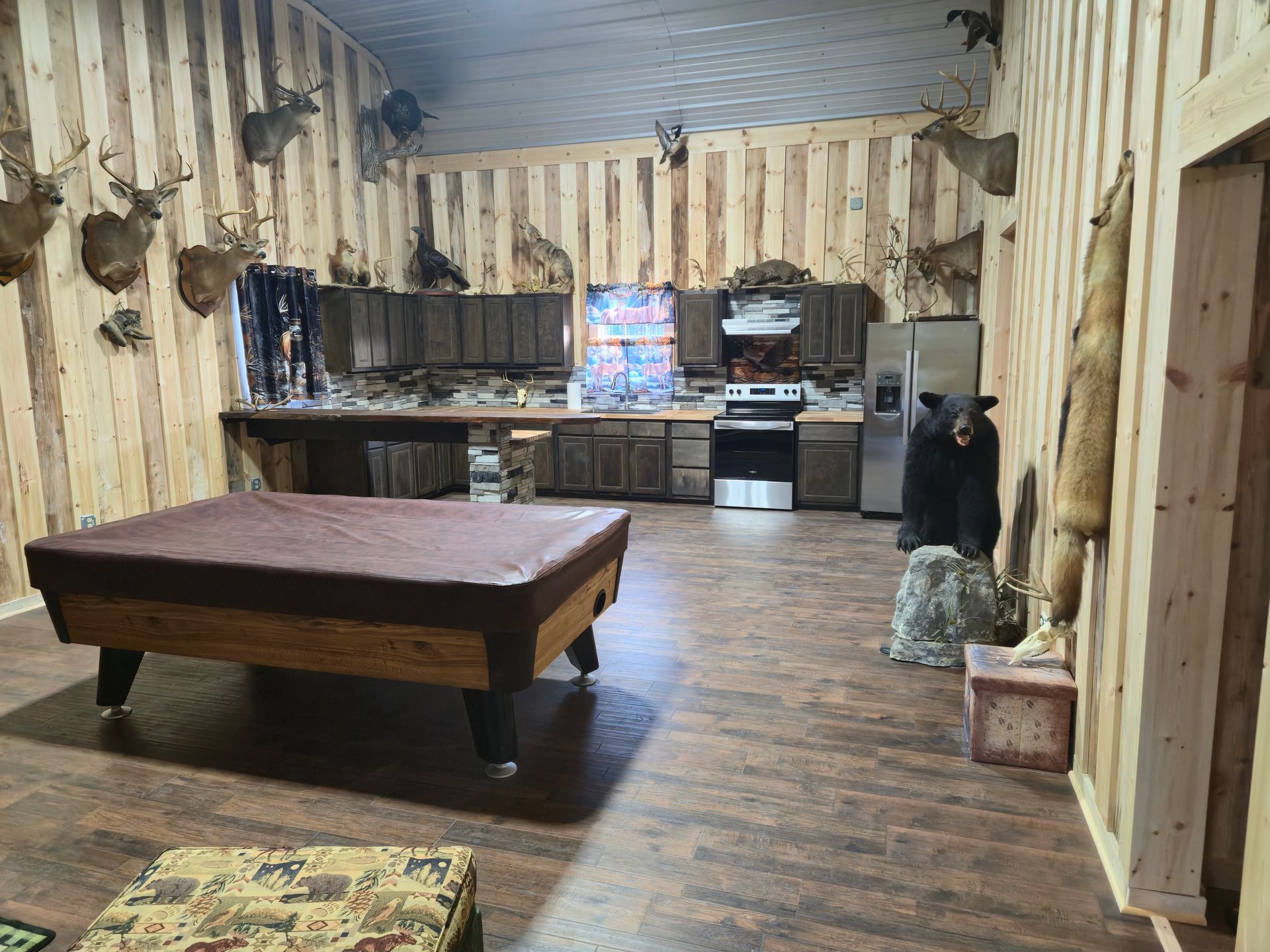 Rustic living room with wood-paneled walls, pool table, and small kitchen area in back