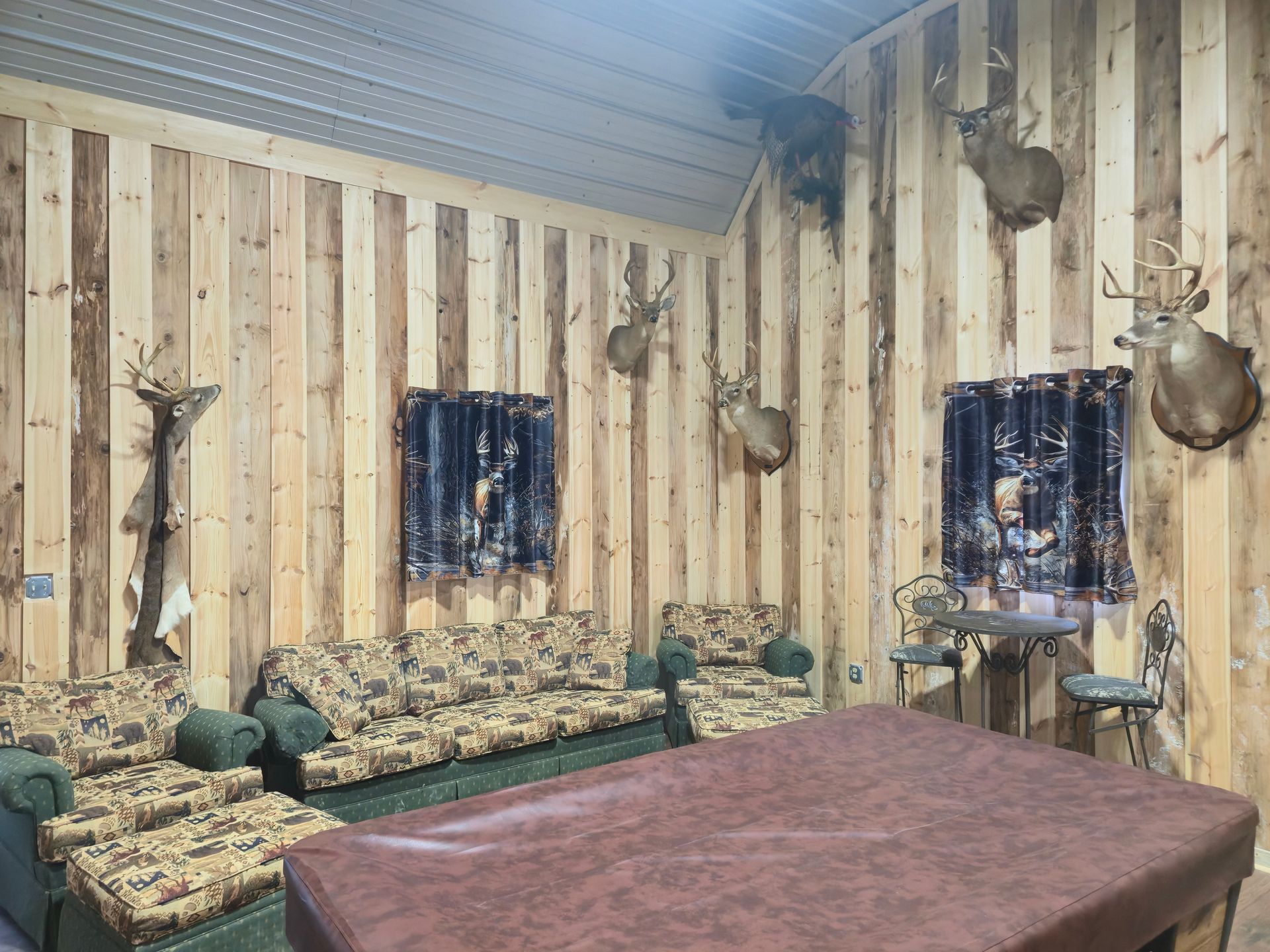Rustic room with wood-paneled walls, mounted antlers, green couches, and a large red table.