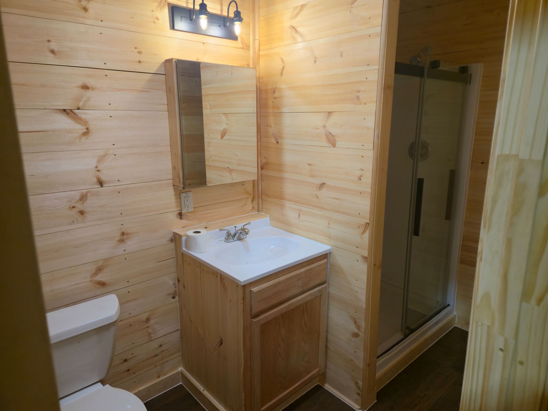 Small wood-paneled bathroom with toilet, sink, mirror, and glass shower stall