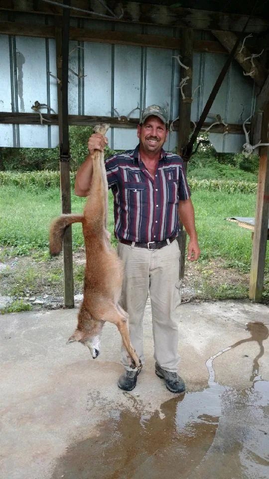 Man holding a large dead fish by the tail in a covered outdoor area, with wet concrete and greenery nearby