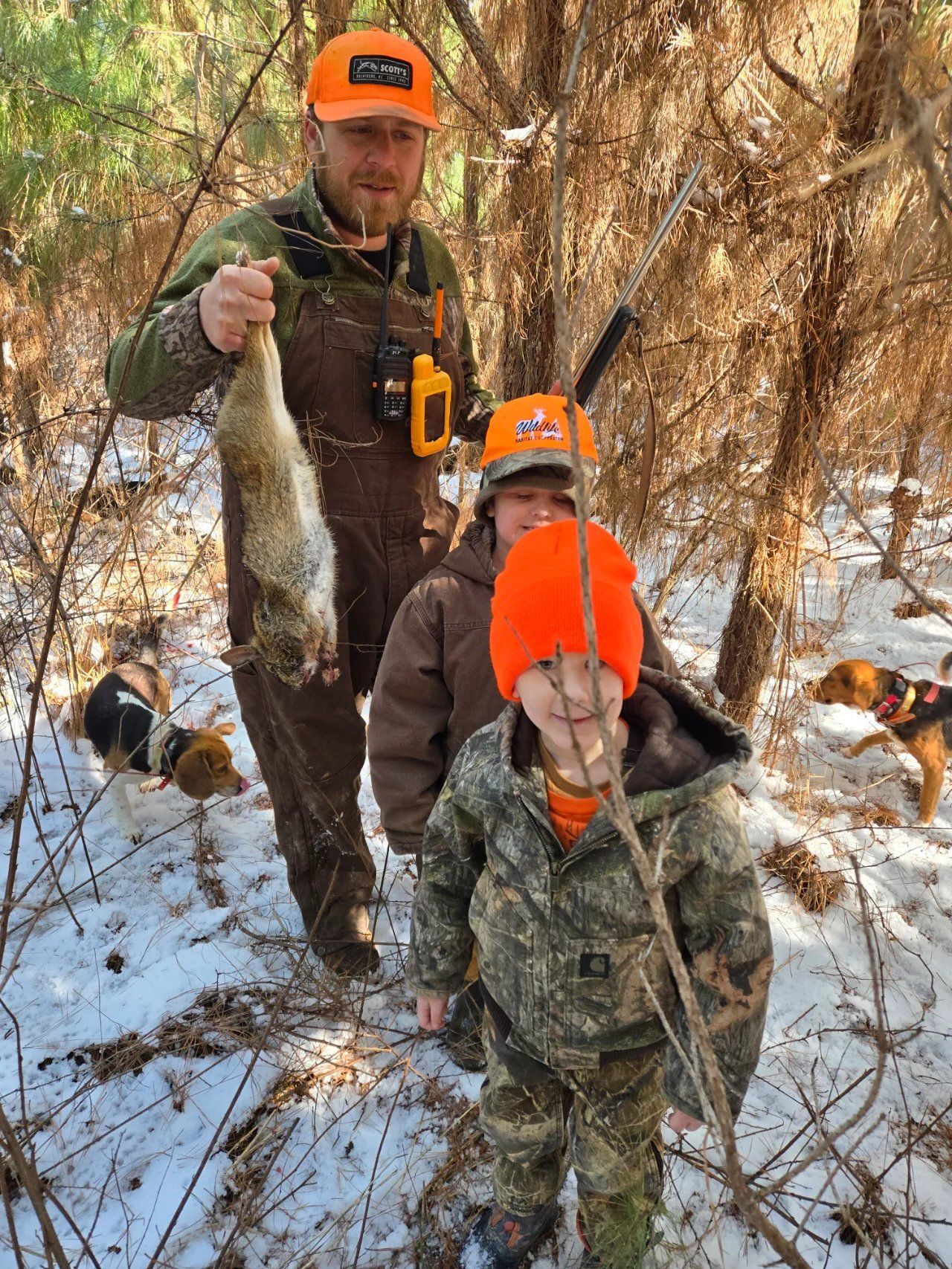 Two people in orange hats pose outdoors in snowy woods, holding a freshly caught fish.