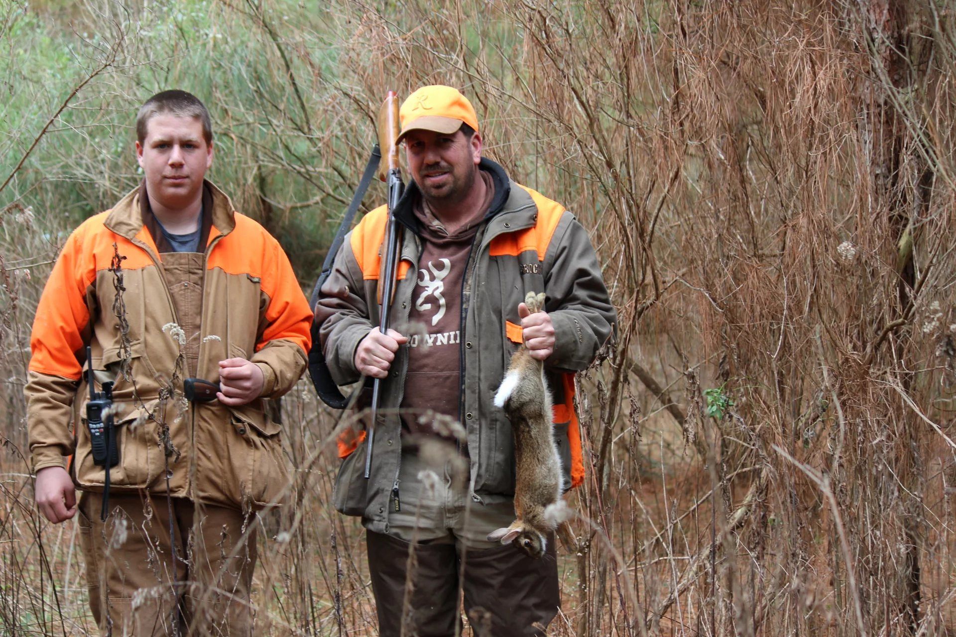 Two hunters in orange gear standing in brush, each holding a rabbit by the ears.