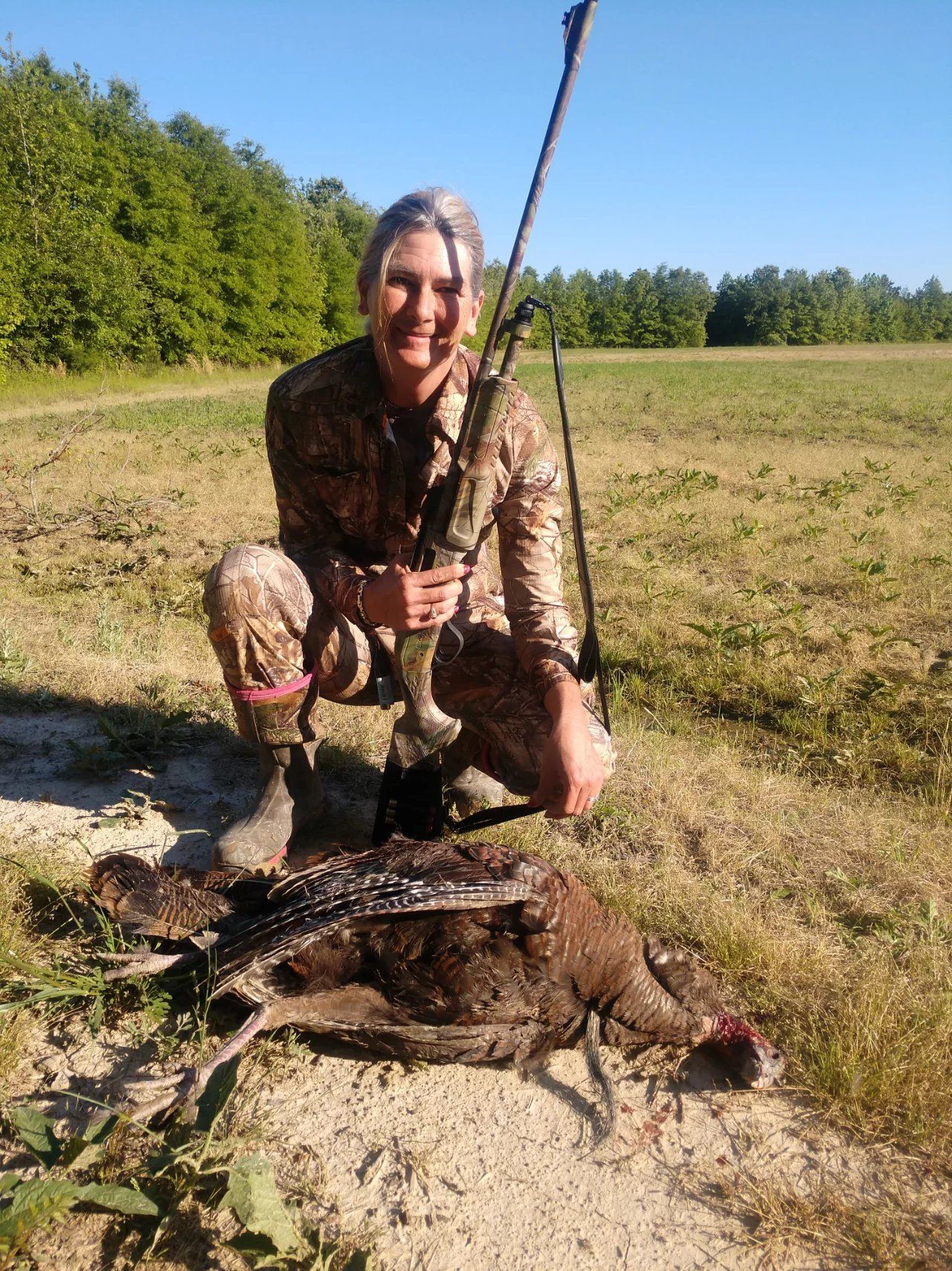 Person kneeling in camouflage beside two hunted animals in a grassy field, holding a rifle.