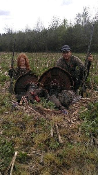 Two people in a field pose with two wild turkeys after a hunt, holding the birds by their tails.