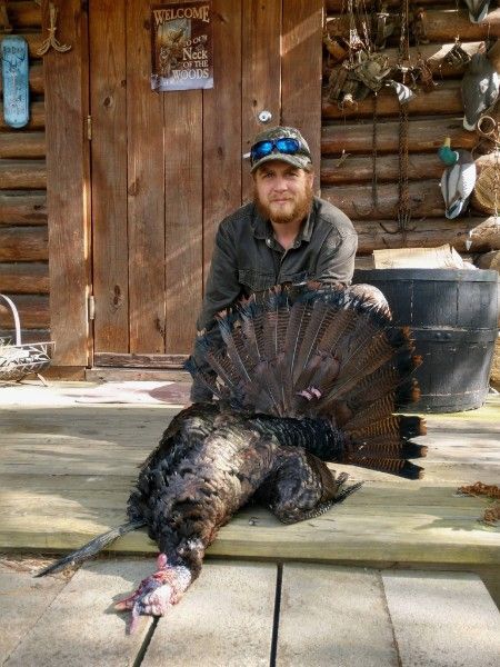 Man posing behind a large dead wild turkey on a wooden porch in front of a cabin
