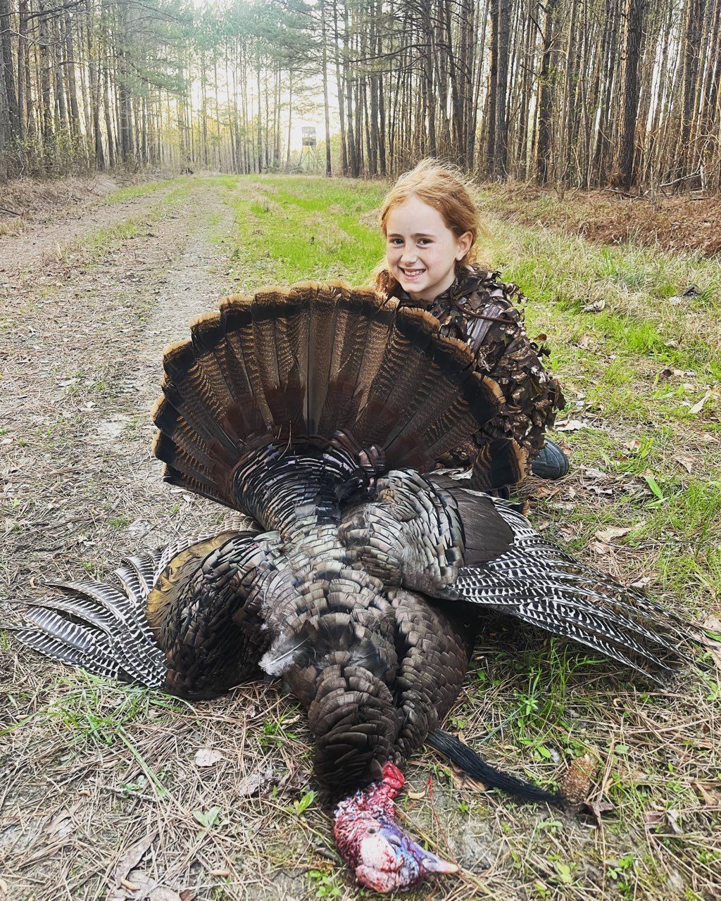 Woman kneeling behind a large wild turkey in a forest clearing, smiling at the camera.