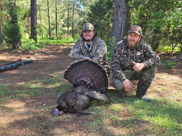 Two hunters in camouflage kneel beside a wild turkey in a wooded clearing.
