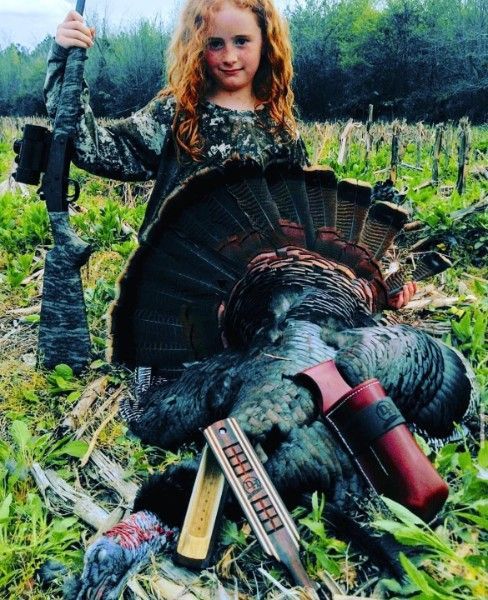 Young child holding a rifle beside a large wild turkey in a field