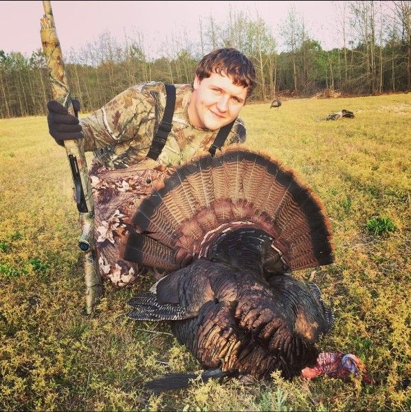 Hunter kneels beside a wild turkey in a grassy field, holding a shotgun at sunset.