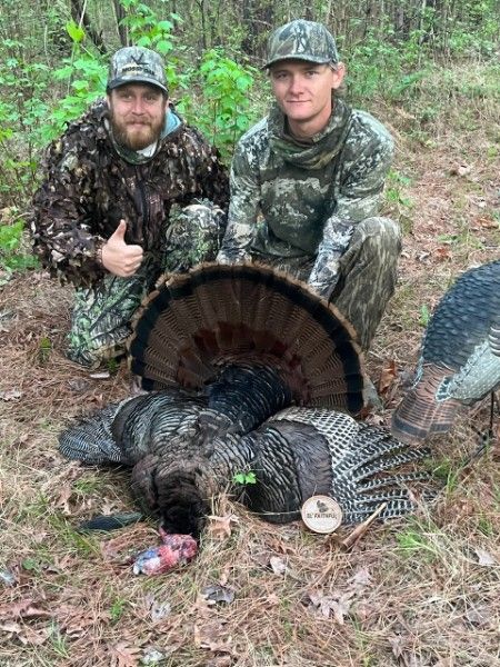Two hunters kneel beside a wild turkey in a wooded area, one giving a thumbs-up.