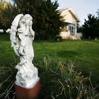 A white angel statue with hands covering its face stands in a yard before a white house with a porch.
