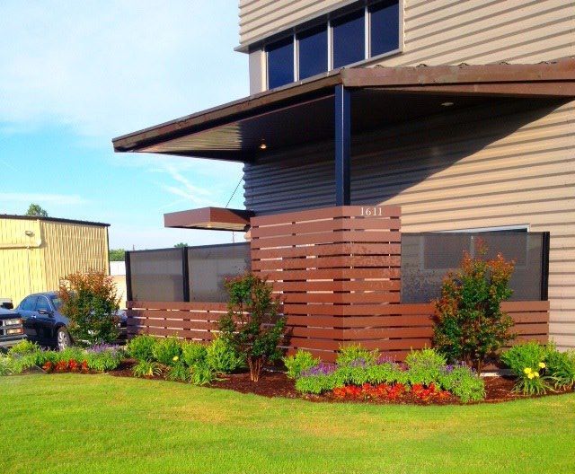 Modern building exterior with a horizontal brown privacy screen, landscaping, and a black metal awning.