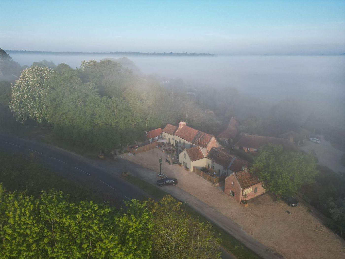 Foggy aerial view of The Barsham Arms pub with red roofs, trees, and a gravel driveway near open water