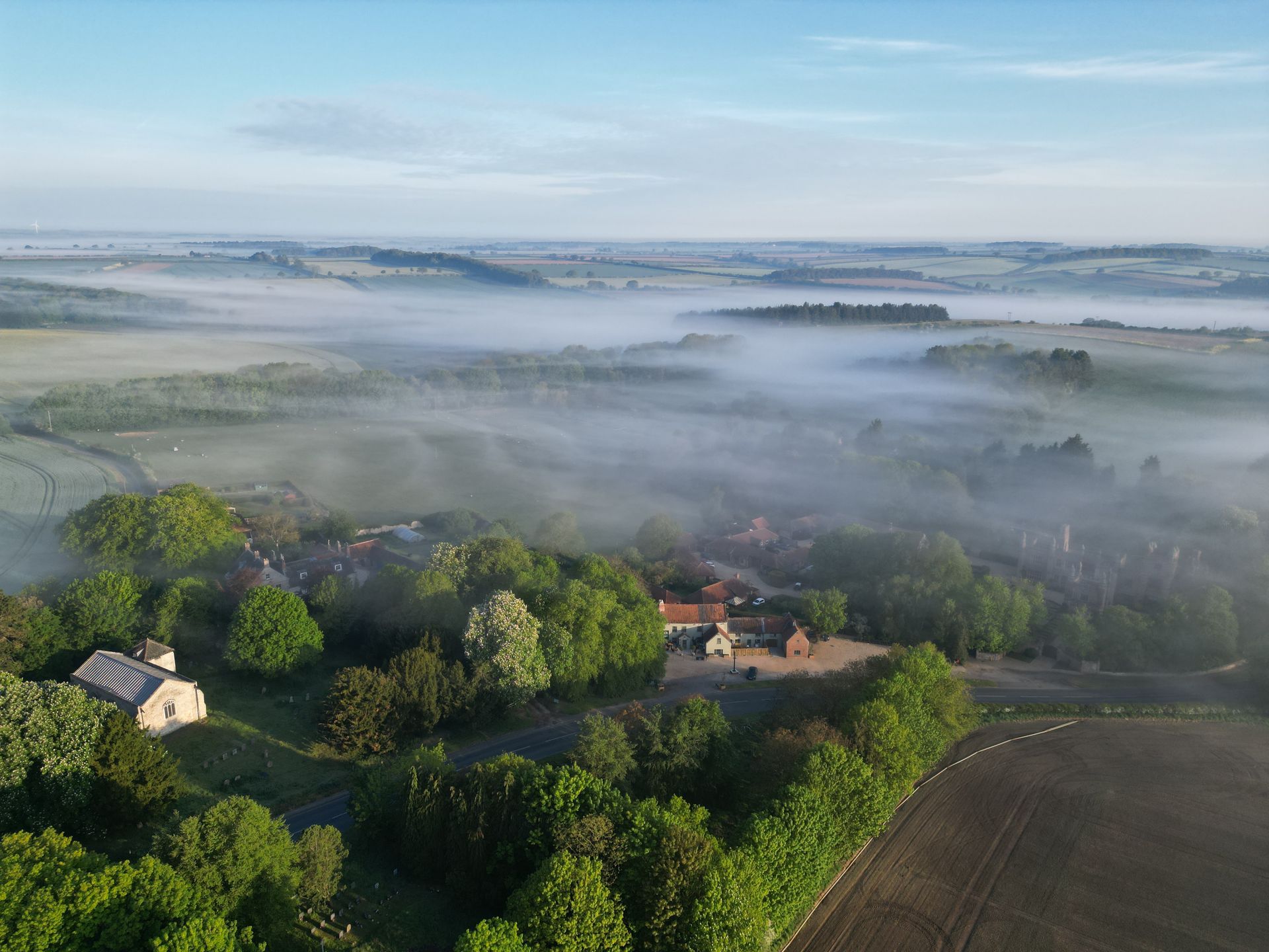 Foggy rural landscape with green fields, scattered houses, and distant hills at sunrise