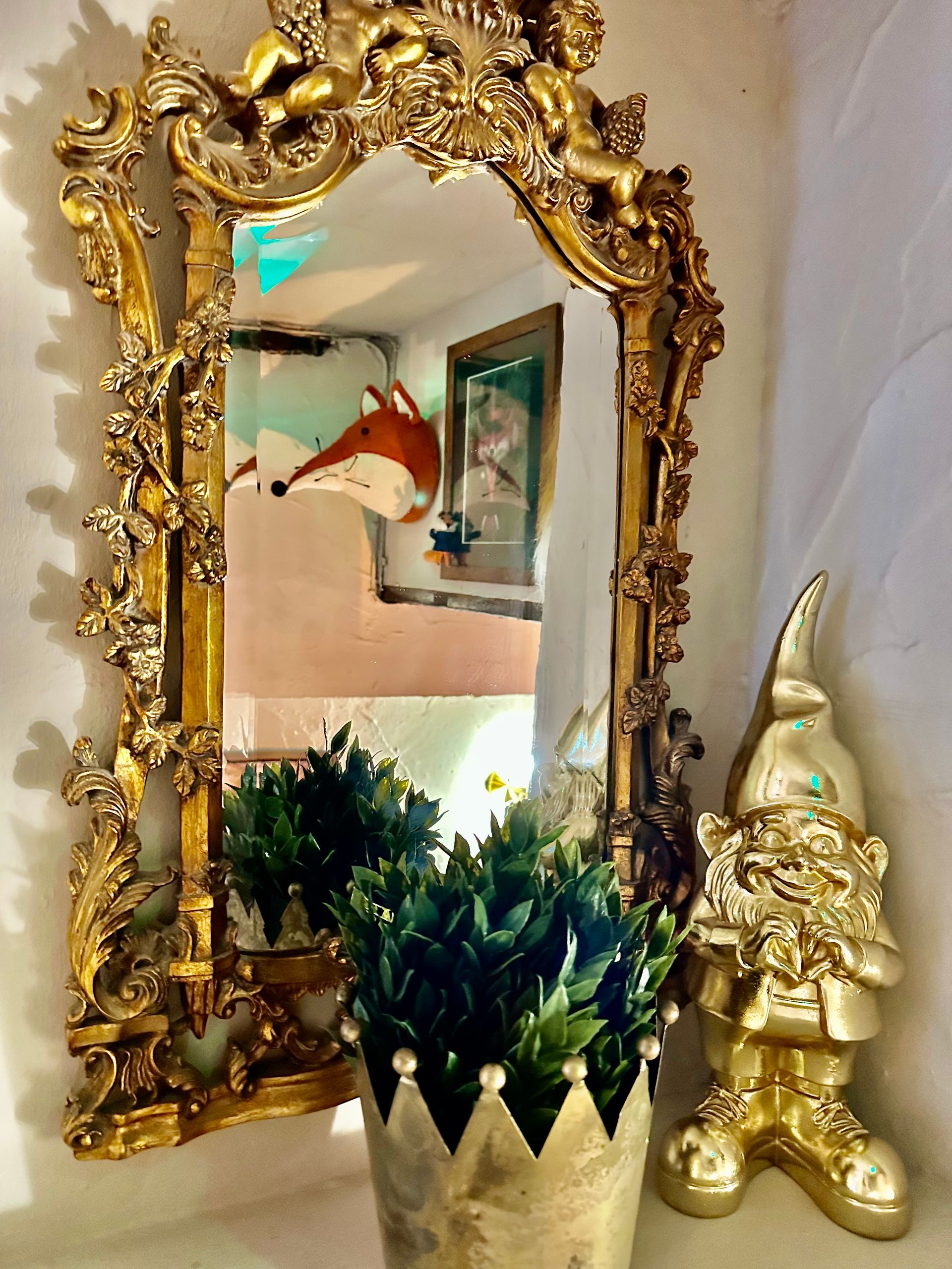 Ornate gold-framed mirror with plant reflection and a small gold gnome figurine beside it