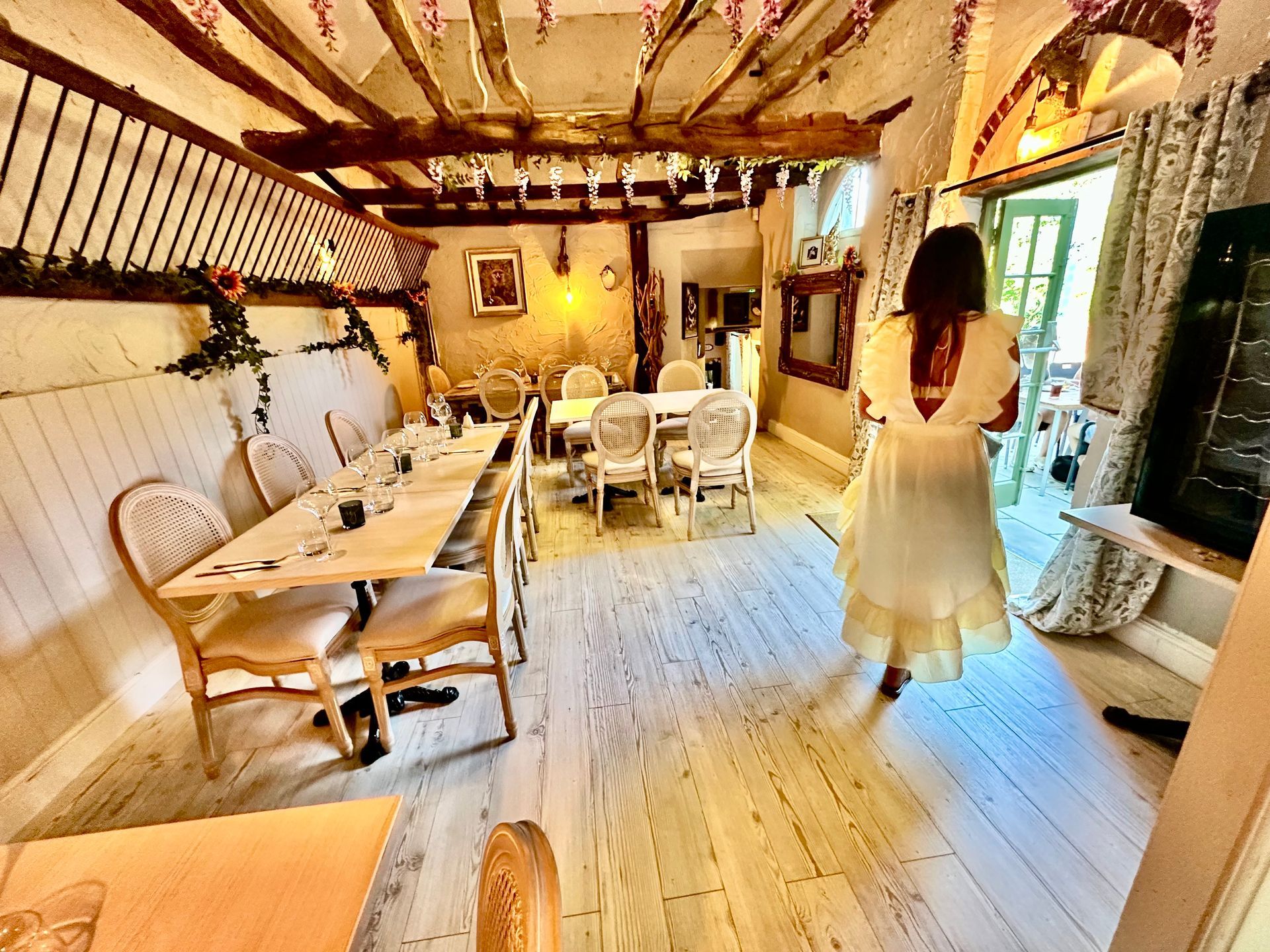 Warm rustic dining room with long table, wicker chairs, and a woman in a white dress near the window