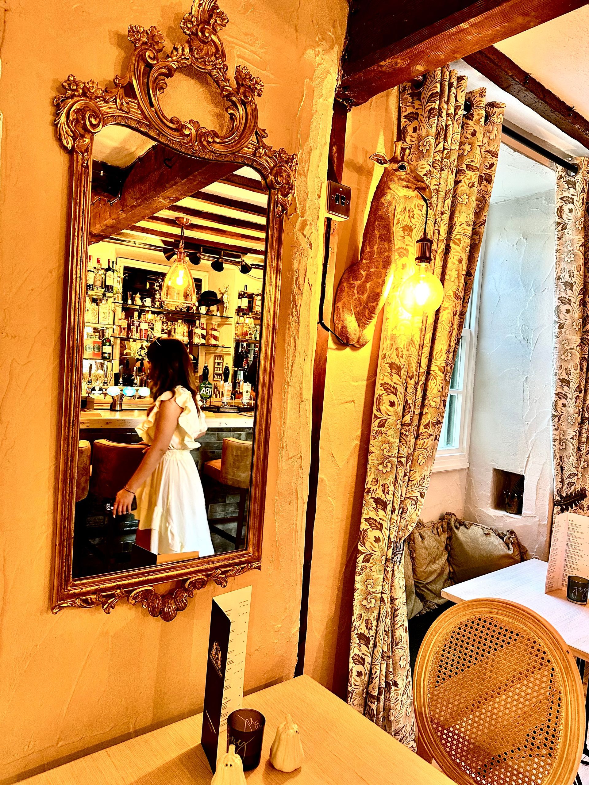 Ornate mirror reflecting a woman in a white dress in a warm, decorative room with drapes and a woven chair