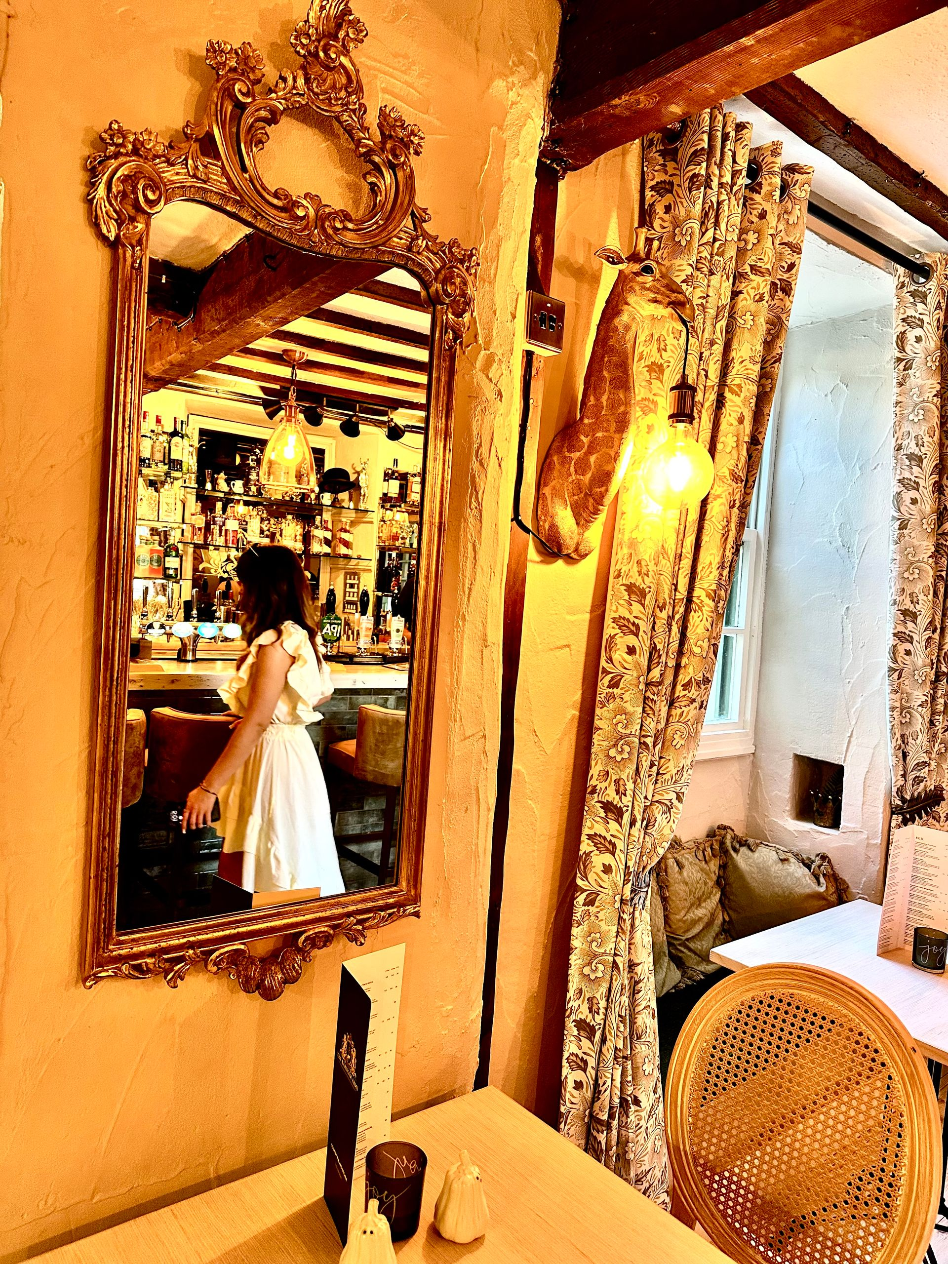 Ornate gold-framed mirror reflecting a chandelier and a person in a white dress in a vintage room