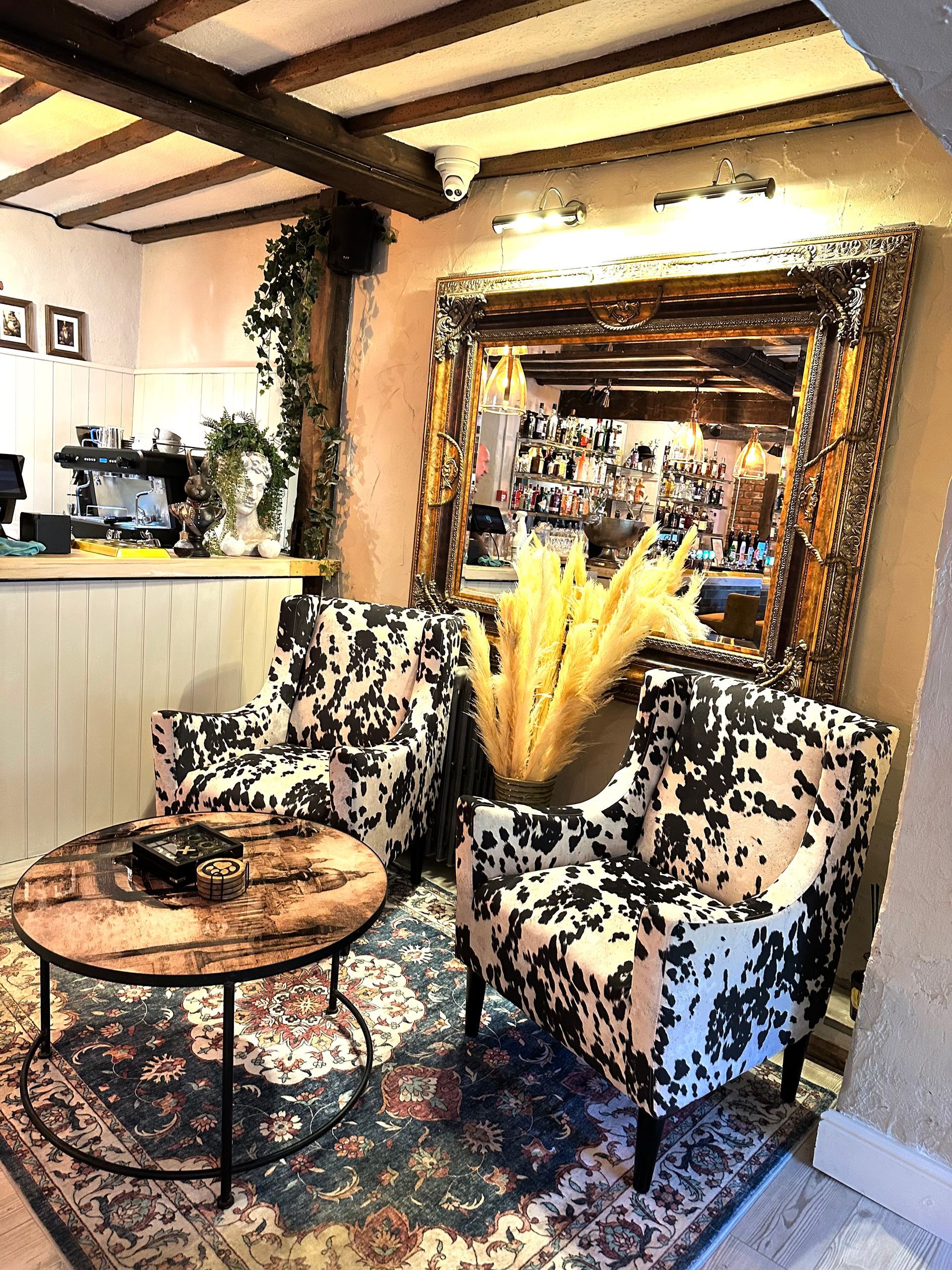 Cozy café seating area with patterned chairs, round table, warm lighting, and a mirror reflecting the room.