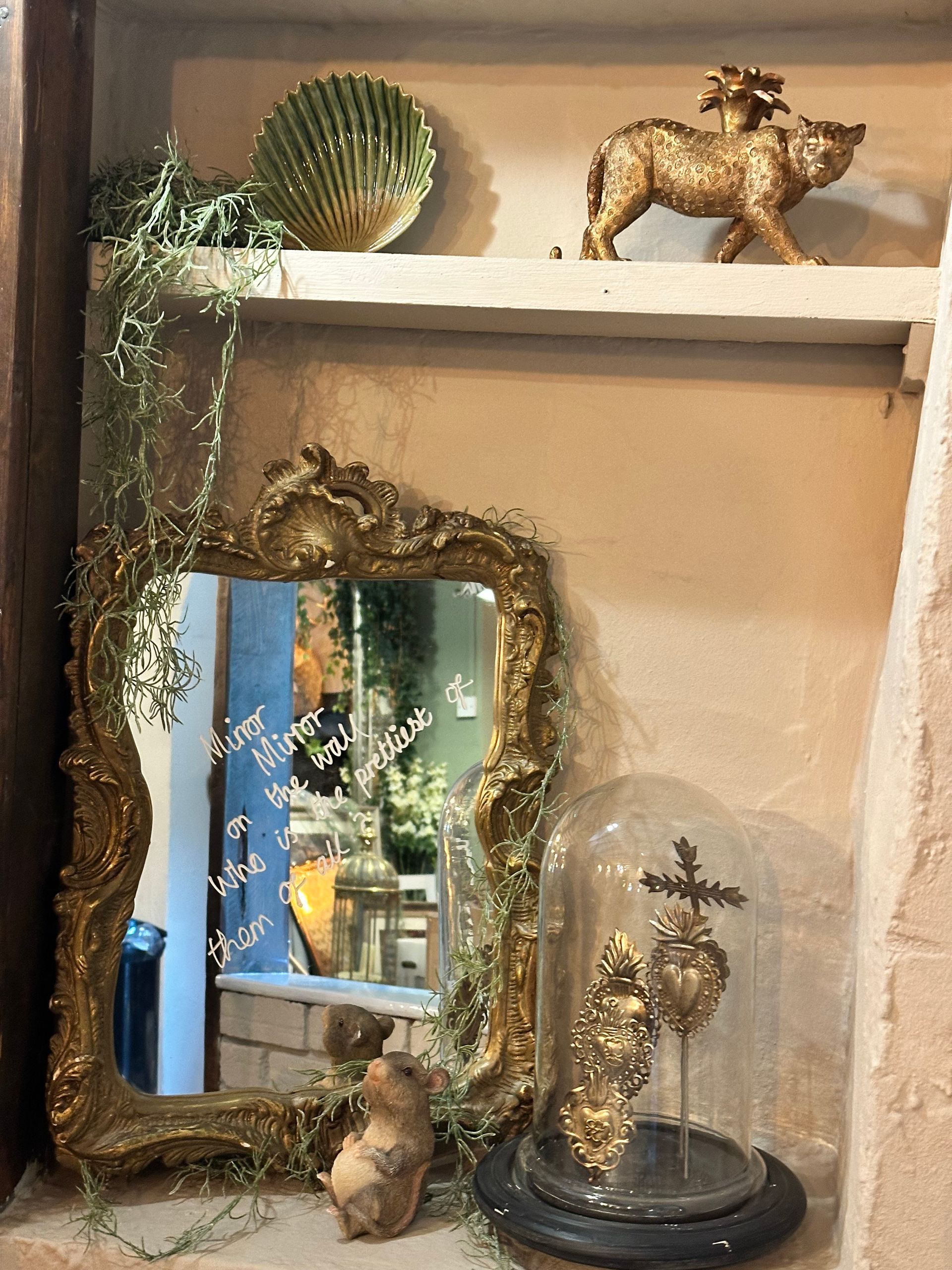 Decorative shelf with gold animal figurines, ornate mirror, and a glass cloche on a light wall with window reflection