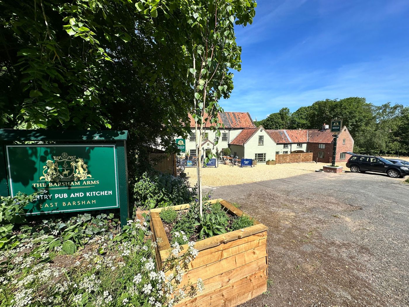 Gravel parking area beside The Barsham Arms pub, with flower beds, trees, and a green signboard.