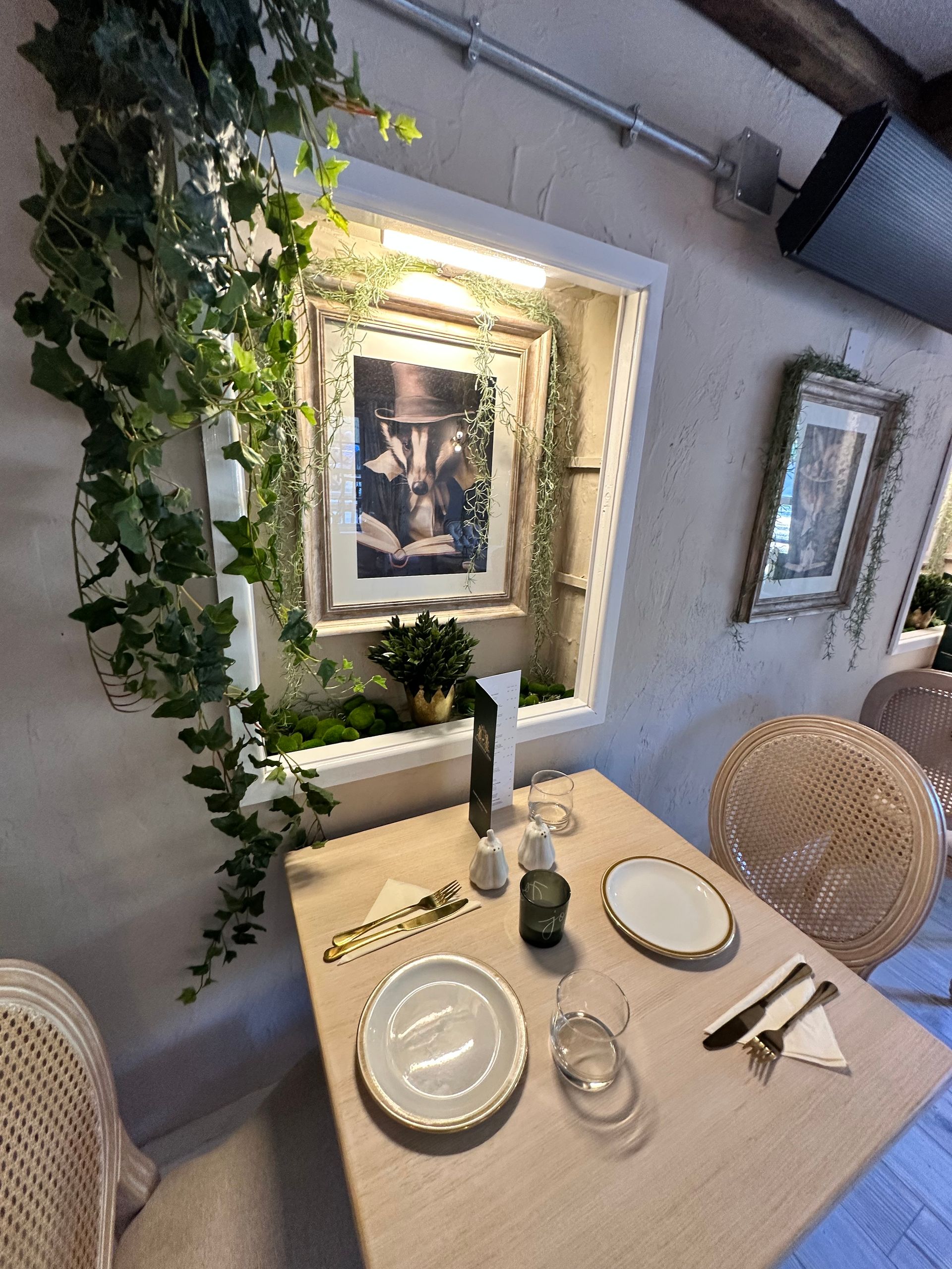 Cozy restaurant table set for two beside a wall niche with framed art and trailing greenery